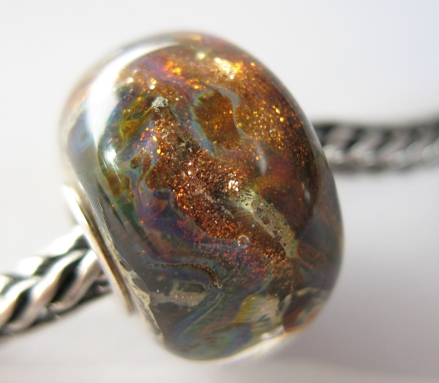 A lovely silver cored lampwork bead with a base of silvered lvory coloured glass with glittery goldstone, purples, greens and blues.